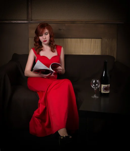 Элегантная молодая рыжая женщина в красном платье, читает гламурный журнал, выпивая бокал красного вина — стоковое фото