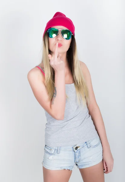 Teenager girl in denim shorts and a gray T-shirt and a pink knit hat, tied at the hips plaid shirt. showing middle finger. girl in sunglasses — Stock Fotó