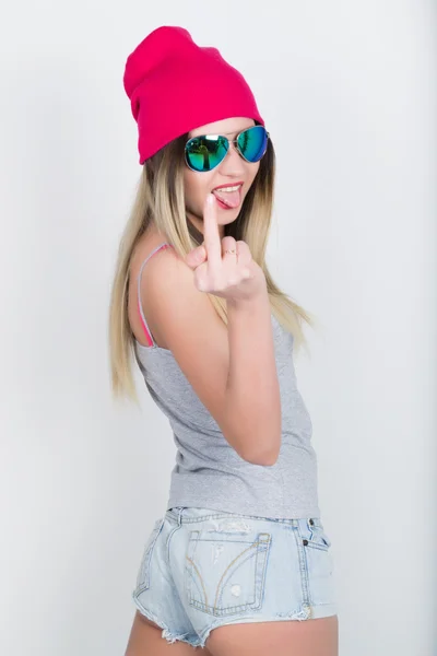 Adolescente en pantalones cortos de mezclilla y una camiseta gris y un sombrero de punto rosa, atado a la camisa a cuadros de las caderas. mostrando el dedo medio. chica en gafas de sol — Foto de Stock