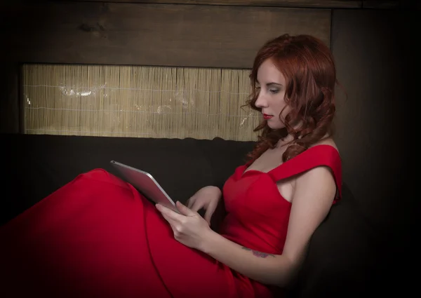 Elegant ung rödhårig kvinna i en röd klänning sitter på soffan och tittar på PC Tablet — Stockfoto