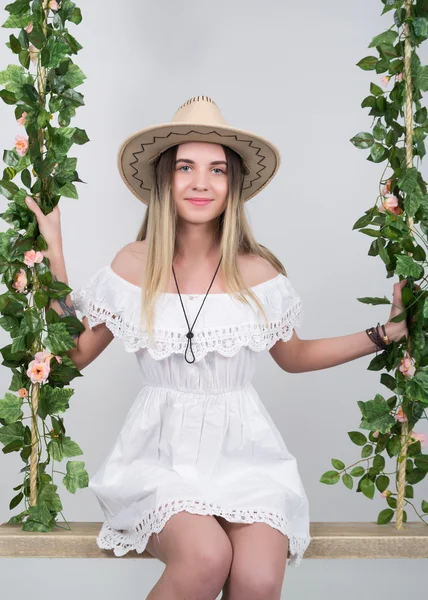 Schöne junge langbeinige Blondine in einem kleinen weißen Kleid und weißem Cowboyhut auf einer Schaukel, hölzerne Schaukel, die an einem Hanfseil hängt, mit Seil umwickelte Weinrebe und Efeu — Stockfoto