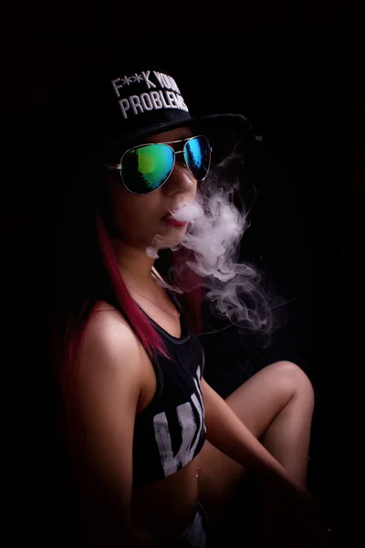 Mujer joven atractiva en gorra y gafas de sol, fuma sobre un fondo oscuro. Ser de moda, ser rapero —  Fotos de Stock