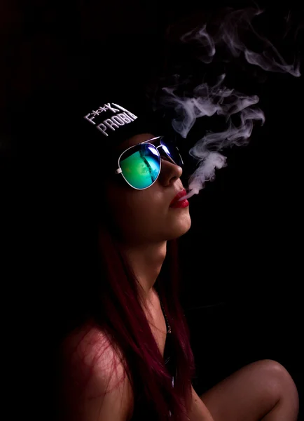 Mujer joven atractiva en gorra y gafas de sol, fuma sobre un fondo oscuro. Ser de moda, ser rapero — Foto de Stock