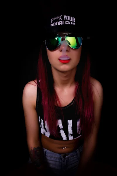 Attractive young woman in cap and sunglasses, smokes on a dark background. Be trendy, be rapper — Stock Photo, Image