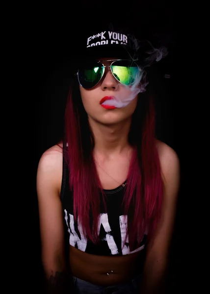 Mujer joven atractiva en gorra y gafas de sol, fuma sobre un fondo oscuro. Ser de moda, ser rapero — Foto de Stock