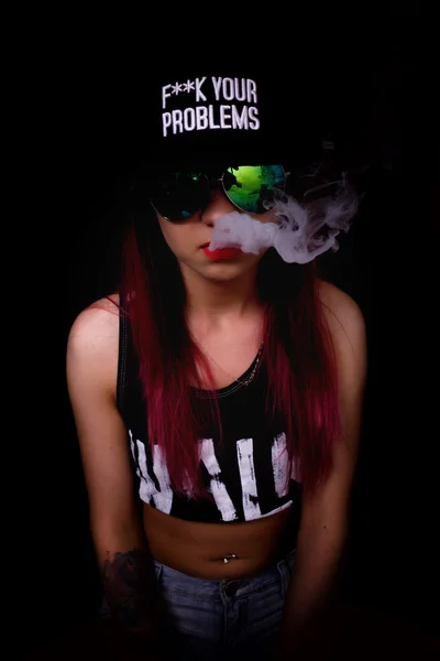 Mujer joven atractiva en gorra y gafas de sol, fuma sobre un fondo oscuro. Ser de moda, ser rapero — Foto de Stock