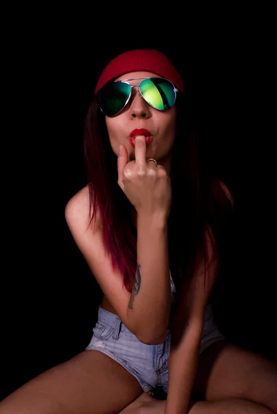 Attractive young woman in a pink knitted cap and sunglasses, sucking her middle finger on a dark background — Stock Photo, Image