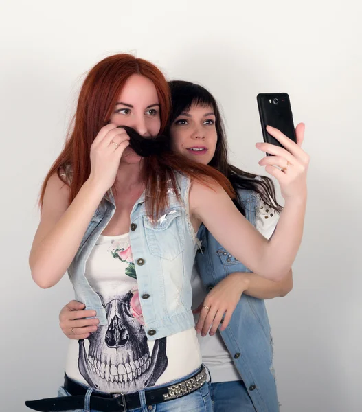Schönheit Hipster Mädchen mit einem Kopfhörer, Selfie auf einem Handy zu machen. Teenager-Fratzen. zwei junge Freundinnen frönen und grimassen, machen einander einen Schnurrbart aus den Haaren — Stockfoto