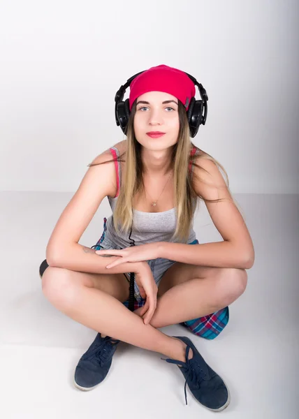 Menina adolescente em calções jeans e uma camiseta cinza e um chapéu de malha rosa, amarrado na camisa xadrez quadris. ele está sentado em um skate e ouvir música em fones de ouvido — Fotografia de Stock