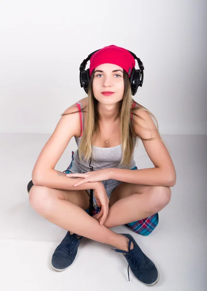 Teenagermädchen in Jeanshosen und grauem T-Shirt und rosa Strickmütze, an den Hüften kariertes Hemd. er sitzt auf einem Skateboard und hört Musik über Kopfhörer — Stockfoto