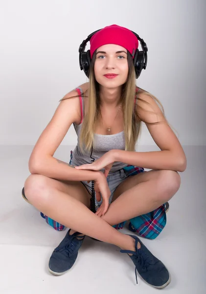 Teenagermädchen in Jeanshosen und grauem T-Shirt und rosa Strickmütze, an den Hüften kariertes Hemd. er sitzt auf einem Skateboard und hört Musik über Kopfhörer — Stockfoto