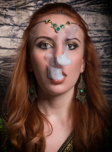 Portrait en gros plan d'une élégante jeune femme rousse, avec maquillage oriental et châle au-dessus de sa tête lèvres rouge vif, fume un narguilé, laisse sortir la fumée de sa bouche — Photo