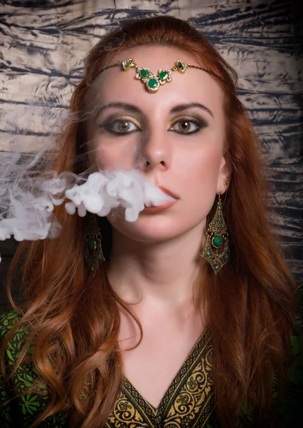 Närbild porträtt av en elegant ung rödhårig kvinna, med orientaliska make-up och sjal över hennes huvud ljusa röda läppar, röker vattenpipa, låt röken ur munnen — Stockfoto