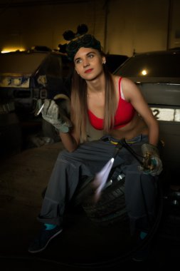 Girl mechanic sitting on a tire with blowpipe smoking in a garage