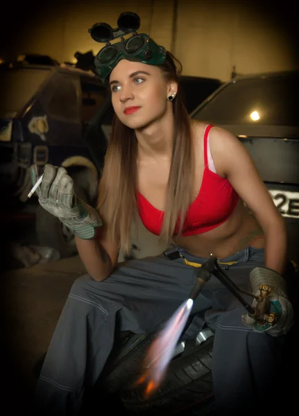 Girl mechanic sitting on a tire with blowpipe smoking in a garage — Stock fotografie