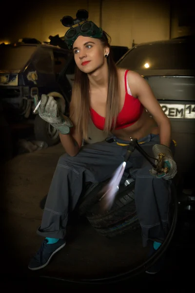 Girl mechanic sitting on a tire with blowpipe smoking in a garage — Φωτογραφία Αρχείου