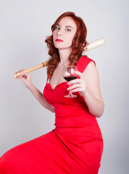 Elegáns fiatal vöröshajú nő egy piros ruhás, vörös bort ittak, és a gazdaság egy baseball ütővel közeli portréja — Stock Fotó