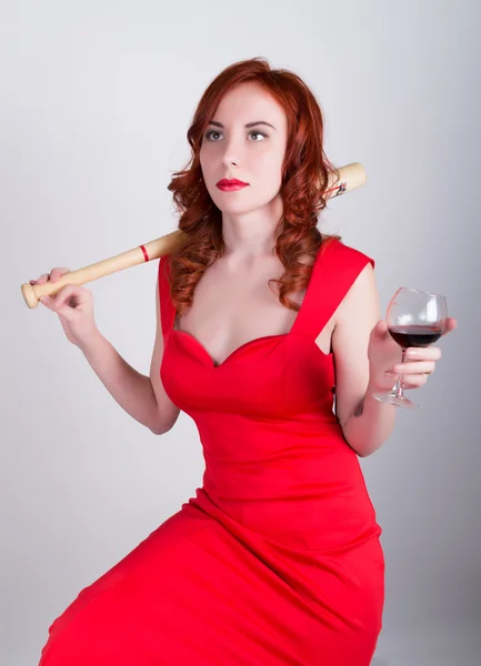 Крупный план портрета Элегантной молодой рыжей женщины в красном платье, пьющей красное вино и держащей бейсбольную биту — стоковое фото