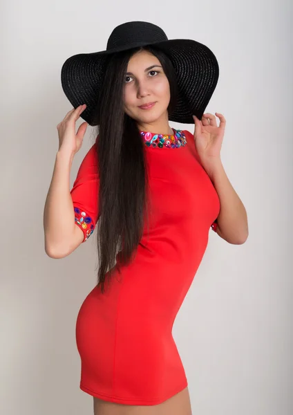 Beautiful asian young brunette in a little red dress and big black hat, standing and posing — Stock Photo, Image