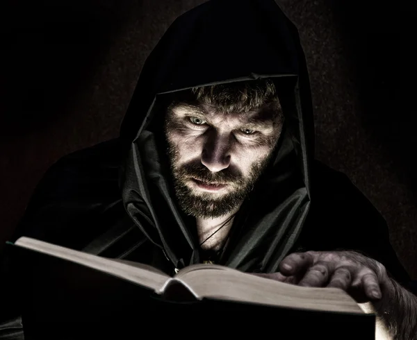 Necromancer werpt spreuken uit dikke oude boek bij kaarslicht op een donkere achtergrond — Stockfoto