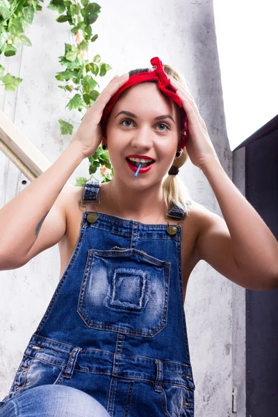 Hermosa mujer joven en overoles de mezclilla y un pañuelo mirando a la cámara como el espejo, Chupa Chups lamiendo y posando — Foto de Stock