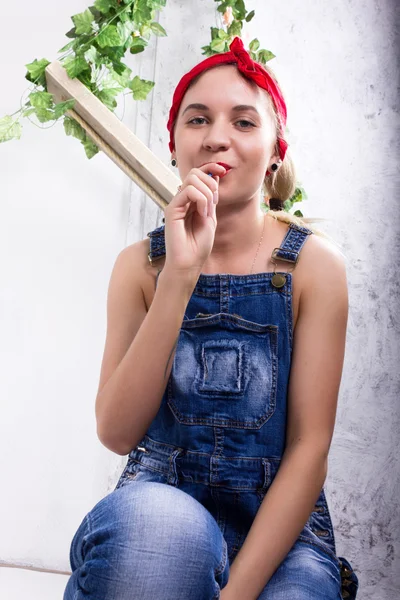 Hermosa mujer joven en overoles de mezclilla y un pañuelo mirando a la cámara como el espejo, Chupa Chups lamiendo y posando — Foto de Stock