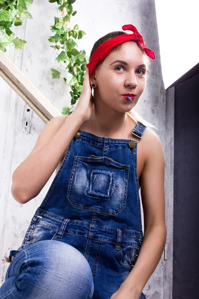 Schöne junge Frau in Jeans-Overalls und einem Kopftuch, die in die Kamera als Spiegel schaut, chupa chups lecken und posieren — Stockfoto