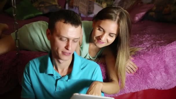 若いカップルは、デジタル タブレット コンピューターを使用してオンライン ショッピング。笑いながら、自宅のベッドや床に横に座っている男の上に横たわる少女のカップル — ストック動画