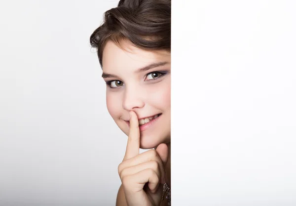 Smiling happy young woman standing behind and leaning on a white blank billboard or placard, expresses different — Stock Photo, Image