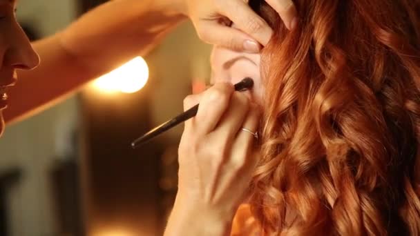 Tijd in een schoonheidssalon. roodharige jonge vrouw in een schoonheidssalon. Stylist maakt make-up bruid op de huwelijksdag. cosmetische poeder borstel voor make-up. — Stockvideo