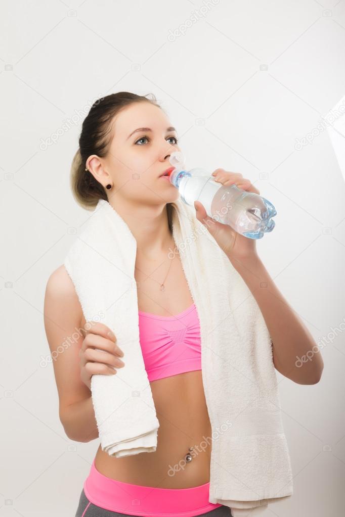 Young athletic girl finished training, holding a bath towel, drinks water from a bottle