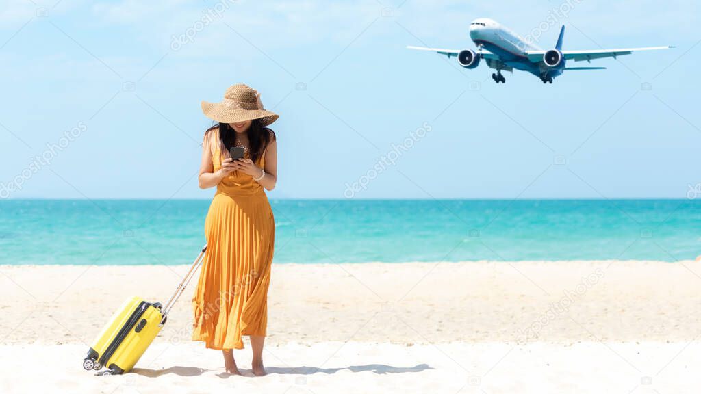 Asia women traveler holding yellow luggage walking and check in smart phone on the beach.  Traveling on an airplane for leisure and destination.  Traveler and Tourism planning trips summer vacations. Summer Concept 