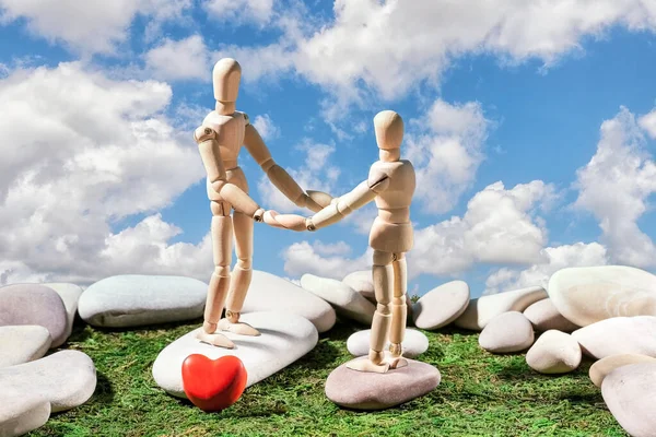 Two mannequins hold hands against a blue sky with white clouds, smooth cobblestones and a heart symbol in the foreground on green grass