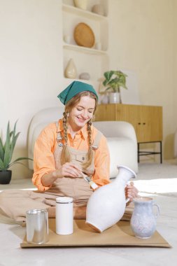 Kadın beyaz tebeşir boyasıyla desenli bir seramik vazo boyuyor, parlak bir oturma odasında DIY 'in sürdürülebilir ev dekorasyonu için yükseldiğini gösteriyor..
