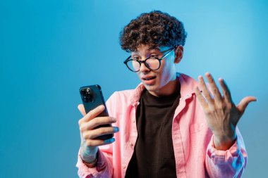 Gözlüklü genç adam video sohbet için akıllı telefon kullanıyor. Mavi stüdyo arka planında sürpriz bir tepki ve modern internet iletişimi gösteriyor..