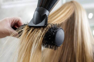 Kurutucu konsantratör ve yuvarlak fırça kullanarak uzun sarı saçlar üzerine profesyonel bir kuaför patlaması sırasında ses ve ses ekleyerek stilistin kapanışı.