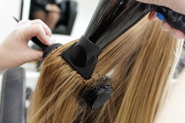 Stilist, profesyonel bir kuaför salonunda uzun sarı saçlara sürtünme sırasında pürüzsüz ve hacim eklemek için yuvarlak fırça ve kurutucu kullanıyor.