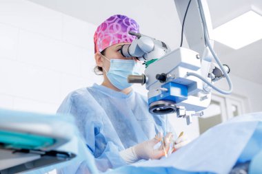 Koruyucu maskeli kadın göz cerrahı. Parlak hastanede vizyon kurtarma prosedürü sırasında mikroskopla ameliyat ediyor.
