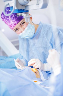 Maskeli gülümseyen kadın cerrah mikroskop altında hassas göz ameliyatları yapıyor.