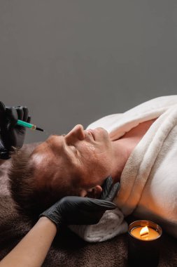 Bornozlu yetişkin bir adam spa 'da yatarken eldivenli bir pratisyen alnına estetik yaptırıyor.