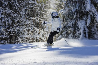 Beyaz ceketli ve kasklı genç kayakçı açık hava sporu konseptiyle karlı orman yamacında hızlı bir dönüş yapıyor..
