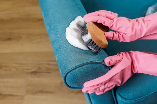 Turkuaz koltuk kolunda köpük fırça kullanan pembe lastik eldivenli eller, uzman döşeme bakımını ve konut lekesi tedavisini gösteriyor.