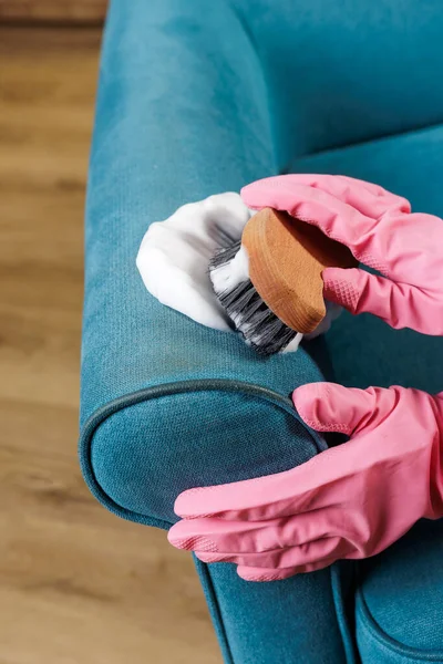 Evdeki döşemelerdeki mavi kumaştan koltuk altlığını temizlemek için köpüklü fırça kullanan pembe lastik eldivenler.