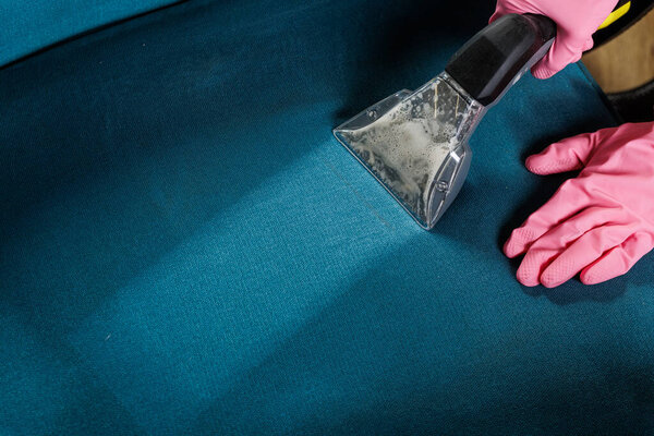 Closeup of cleaner in pink gloves using wet vacuum extractor to wash sofa seat, showing deep upholstery cleaning and stain removal service.