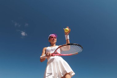 Dişi tenis atleti, tezgahlı mavi gökyüzüne karşı güneş ışığında raketle atış yapmaya hazırlanıyor.