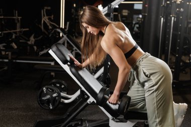 Kadın, spor salonunda forma ve kuvvete odaklanan eğimli bankta desteklenen bir kol dambılı ile kaslarını çalıştırıyor.