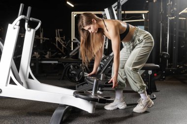 Atletik kadın, modern spor salonundaki direnç antrenmanına hazırlanan fitness makinesine ağırlık tabağı ekliyor.