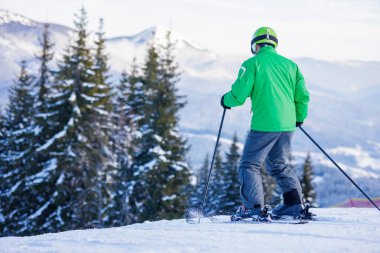 Yeşil ceketli ve kasklı kayakçının kar üzerinde sırık, çam ormanı ve kayak merkezindeki dağ manzaralı görüntüsü