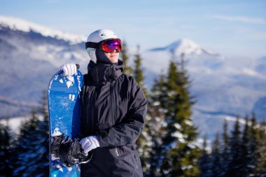 Kasklı ve gözlüklü snowboard binicisi karlı dağda mavi snowboard, çam ormanı ve alp zirveleri taşıyor.