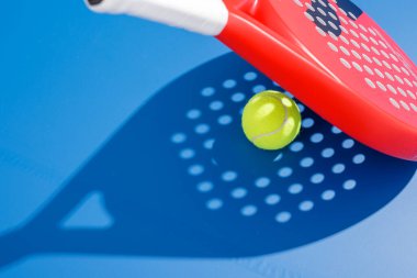 Padel tenis topu raketin yanında süzülüyor ve mavi kortun arka planında pankart tasarımı ve tanıtımı için delinmiş gölge var.